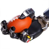 A black robotic hand with five jointed fingers mimicking a human hand holds an orange miniature football.